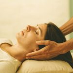 Une femme aux yeux fermés reçoit un massage des tempes par deux mains sur un oreiller.